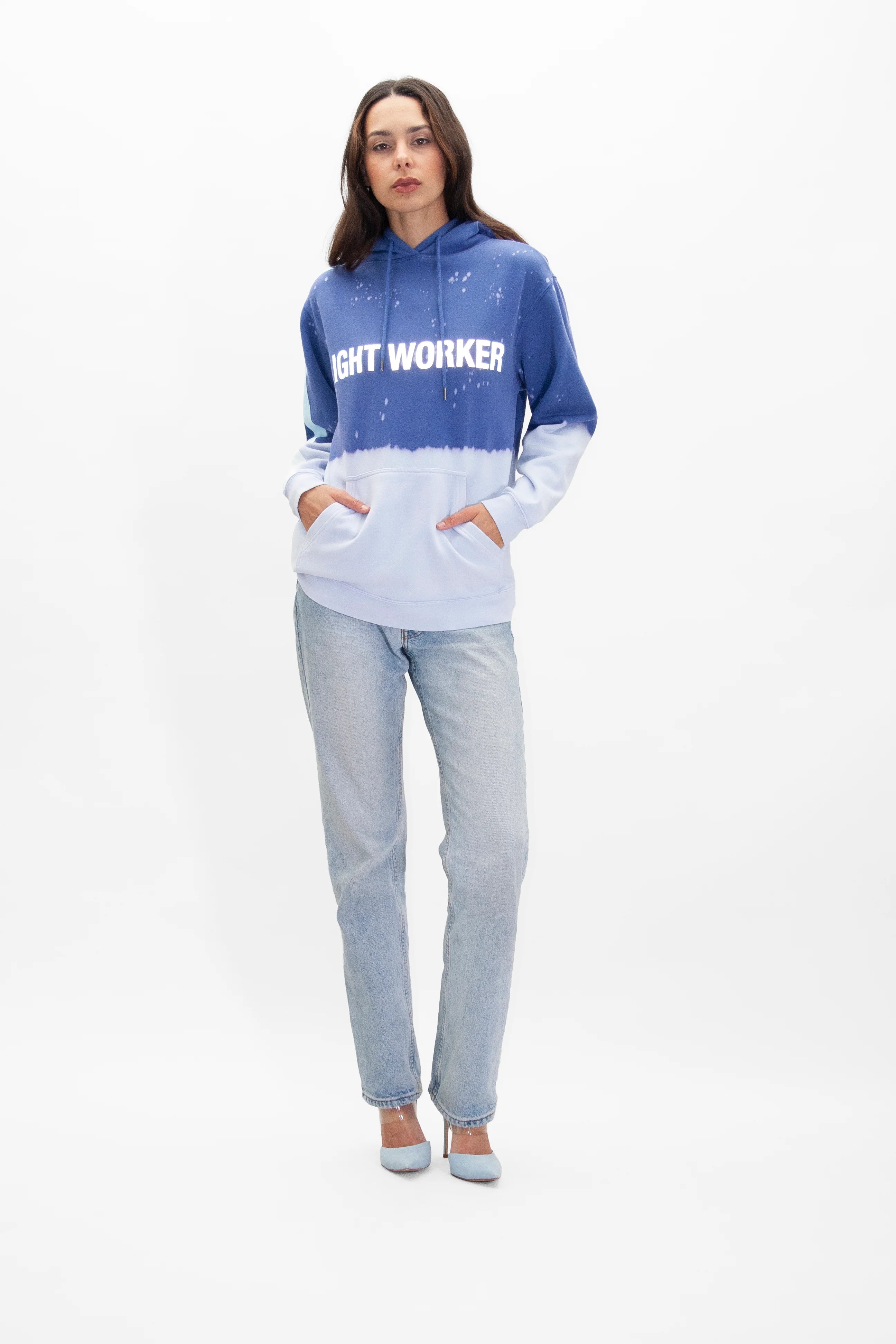 A person stands wearing a Limited Edition Light Worker Hoodie in Indigo Child by GFLApparel, blue jeans, and gray shoes against a plain white background. Their hands are comfortably tucked into the hoodie's pockets, embodying the essence of an Indigo Child.