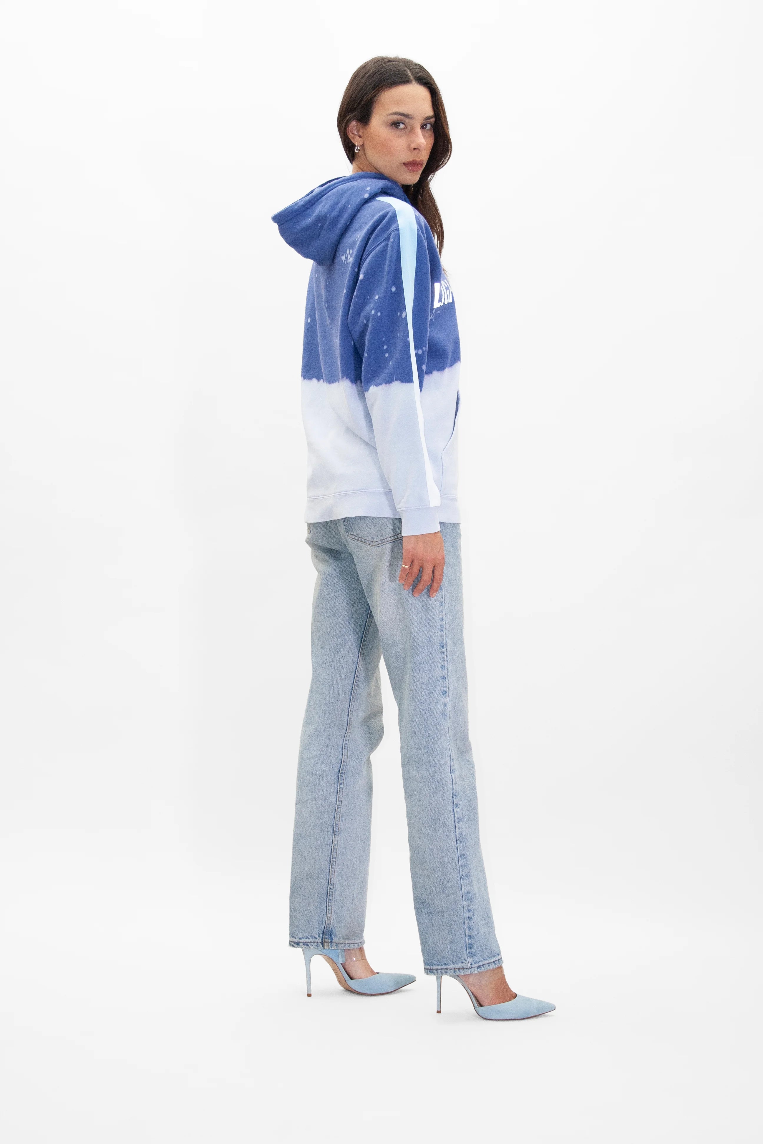 Woman in a blue and white Limited Edition Light Worker Hoodie in Indigo Child by GFLApparel, light blue jeans, and blue high heels, standing and looking back over her shoulder against a plain white background.