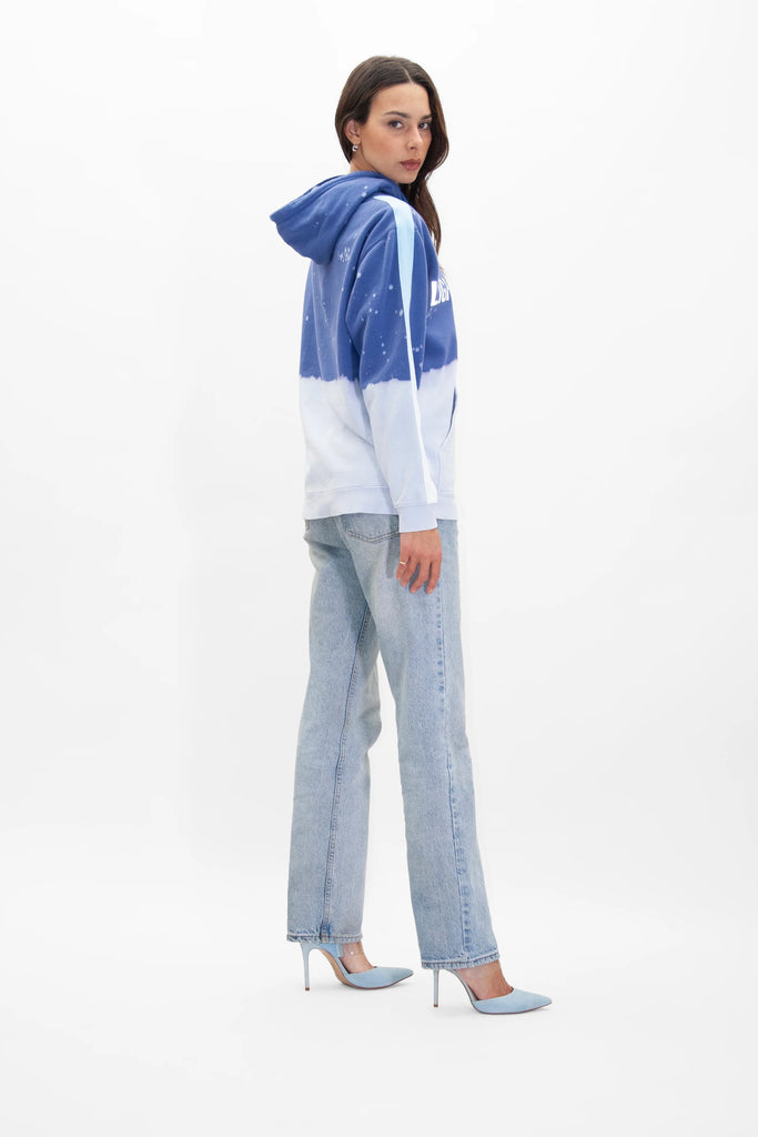 Woman in a blue and white Limited Edition Light Worker Hoodie in Indigo Child by GFLApparel, light blue jeans, and blue high heels, standing and looking back over her shoulder against a plain white background.
