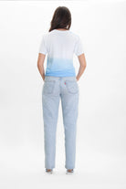 A person with long dark hair stands facing away, wearing a white and blue Light Worker Tee in Atmosphere by GFLApparel featuring an ombré gradient wash, light blue jeans, and white shoes against a plain white background.