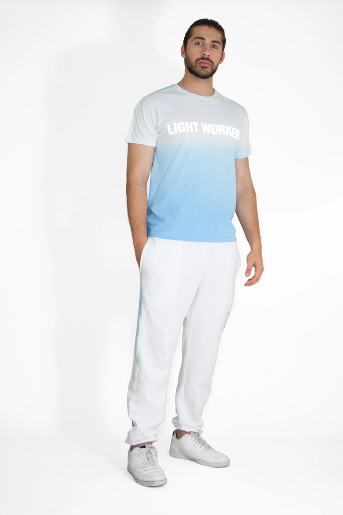 A man wearing a GFLApparel LIGHT WORKER TEE IN ATMOSPHERE t-shirt and white sweatpants.