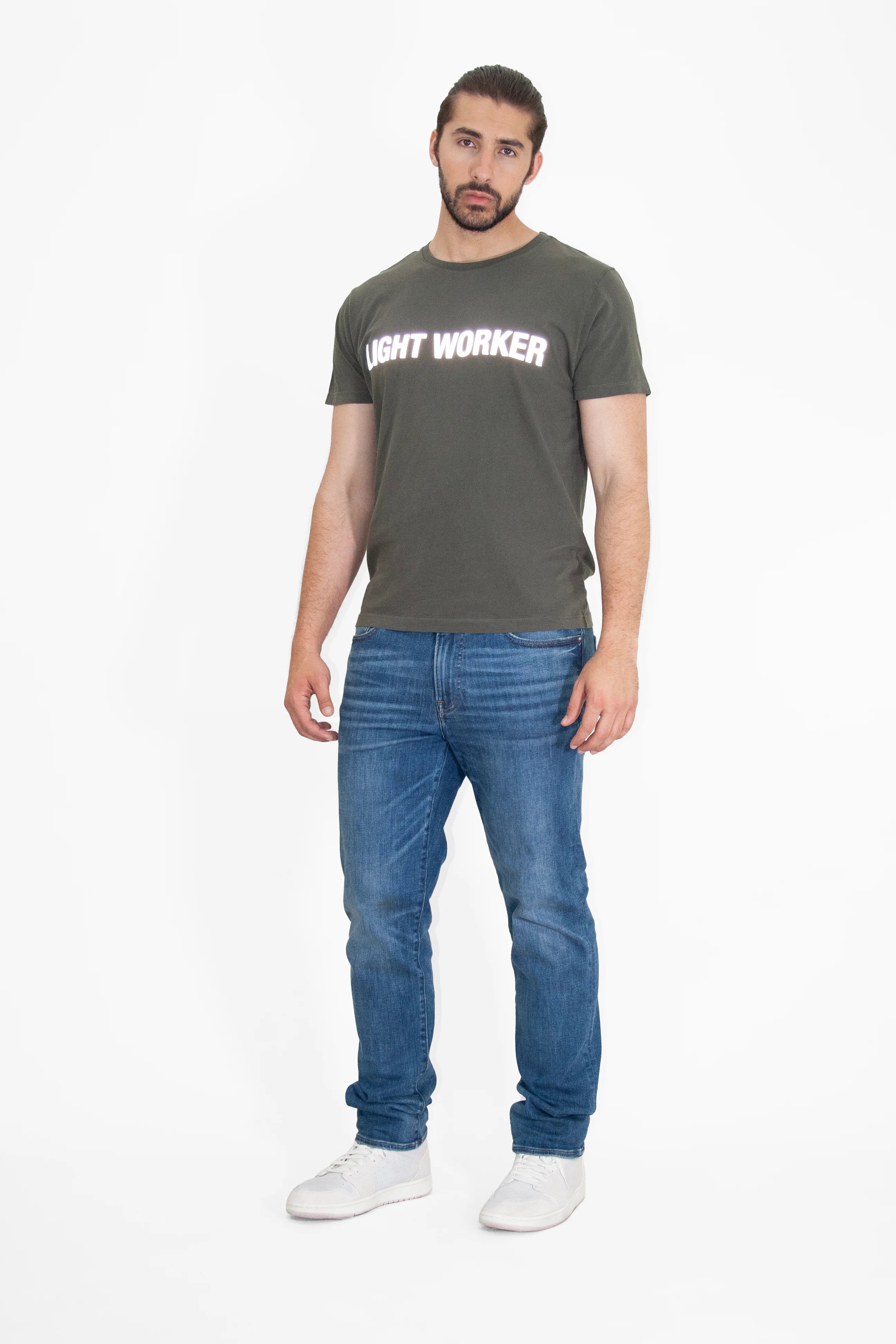 A man with long hair tied back, wearing a dark Light Worker Tee in Caladan from GFLApparel made from breathable cotton, blue jeans, and white sneakers, stands against a plain white background.