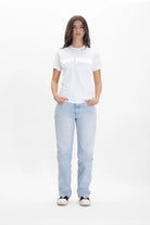 A person with long hair stands casually with hands in pockets, wearing a white t-shirt that says "FIGHT WORKER" in reflective print and light blue jeans. The Light Worker Tee in Lite Beam from GFLApparel is made of soft, breathable cotton. The background is plain white.