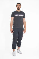 A person stands against a plain white background wearing a black GFLApparel Light Worker Tee in Space Glow made of breathable cotton, black jogger pants, and white sneakers.