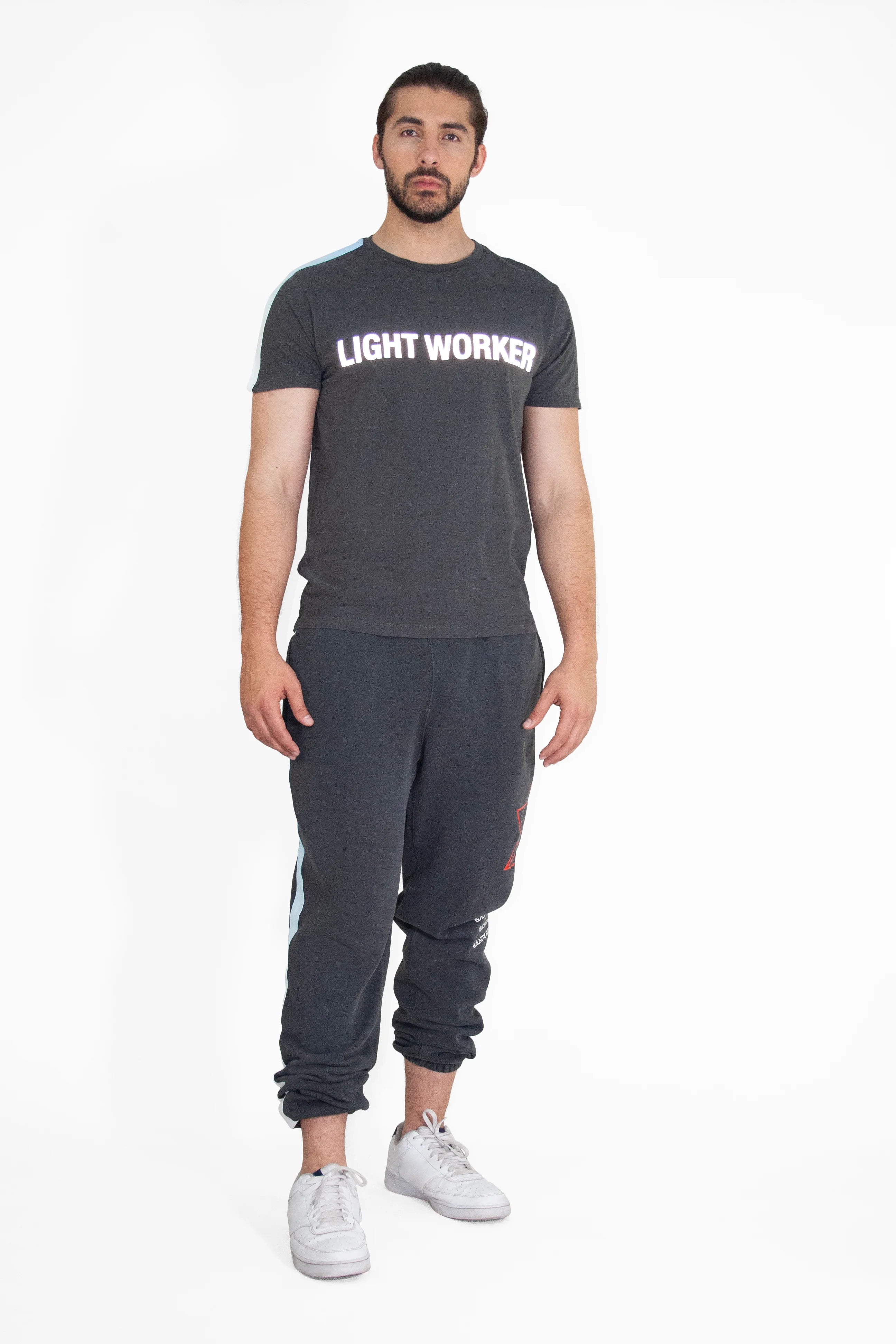 A person stands against a plain white background wearing a black GFLApparel Light Worker Tee in Space Glow made of breathable cotton, black jogger pants, and white sneakers.
