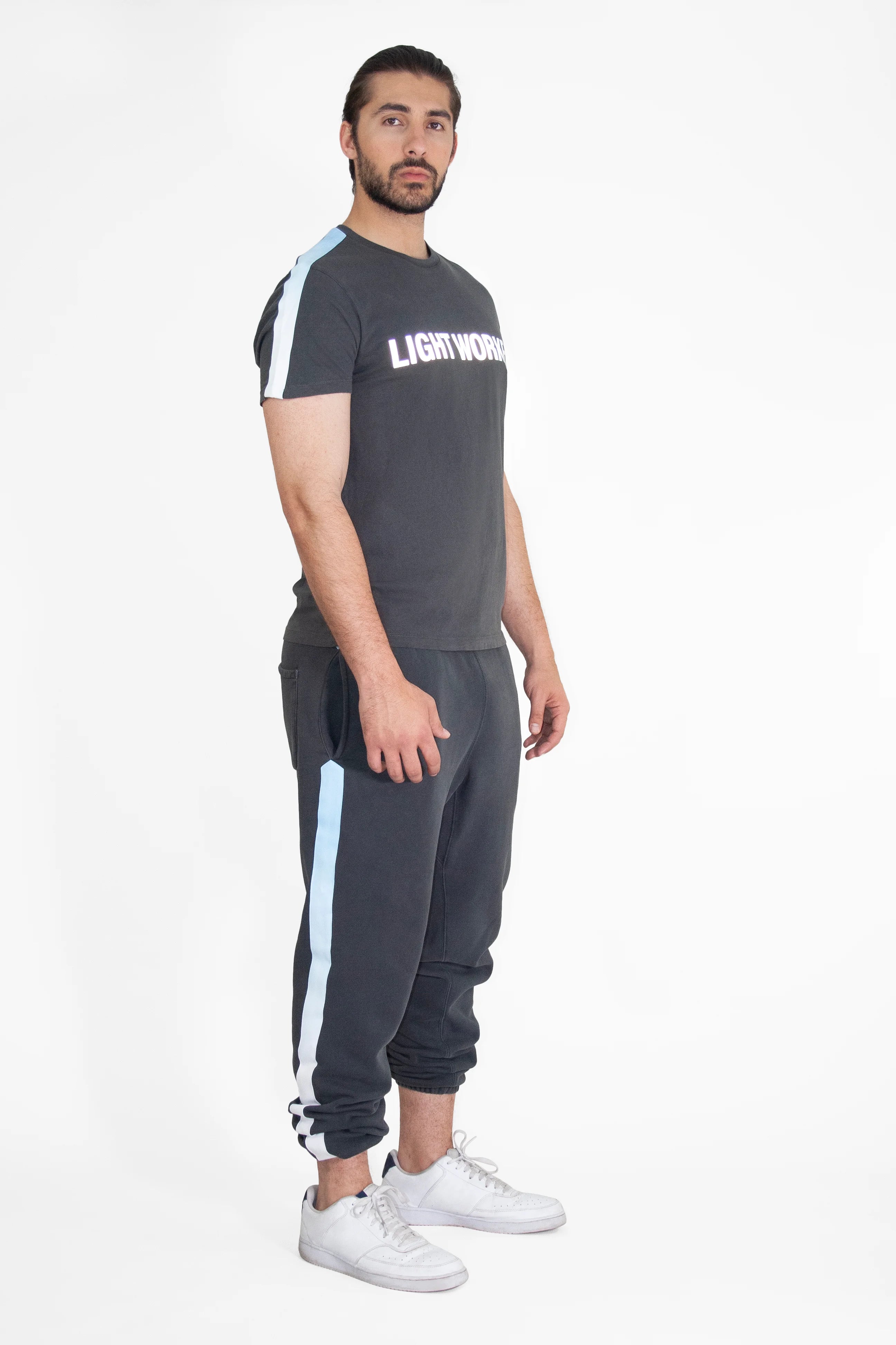 A man with dark hair and beard is standing in profile. He's wearing a black T-shirt with "Light Worker Tee in Space Glow" by GFLApparel reflective print on the front, breathable cotton black joggers with light blue stripes, and white sneakers.