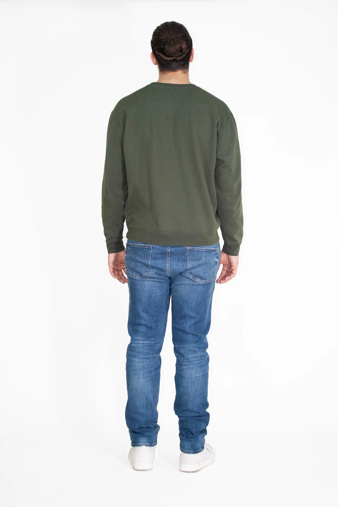 A person with long hair in a bun is standing with their back facing the camera, wearing a GFLApparel Light Worker Crewneck in Caladan, blue jeans, and white shoes against a plain white background.