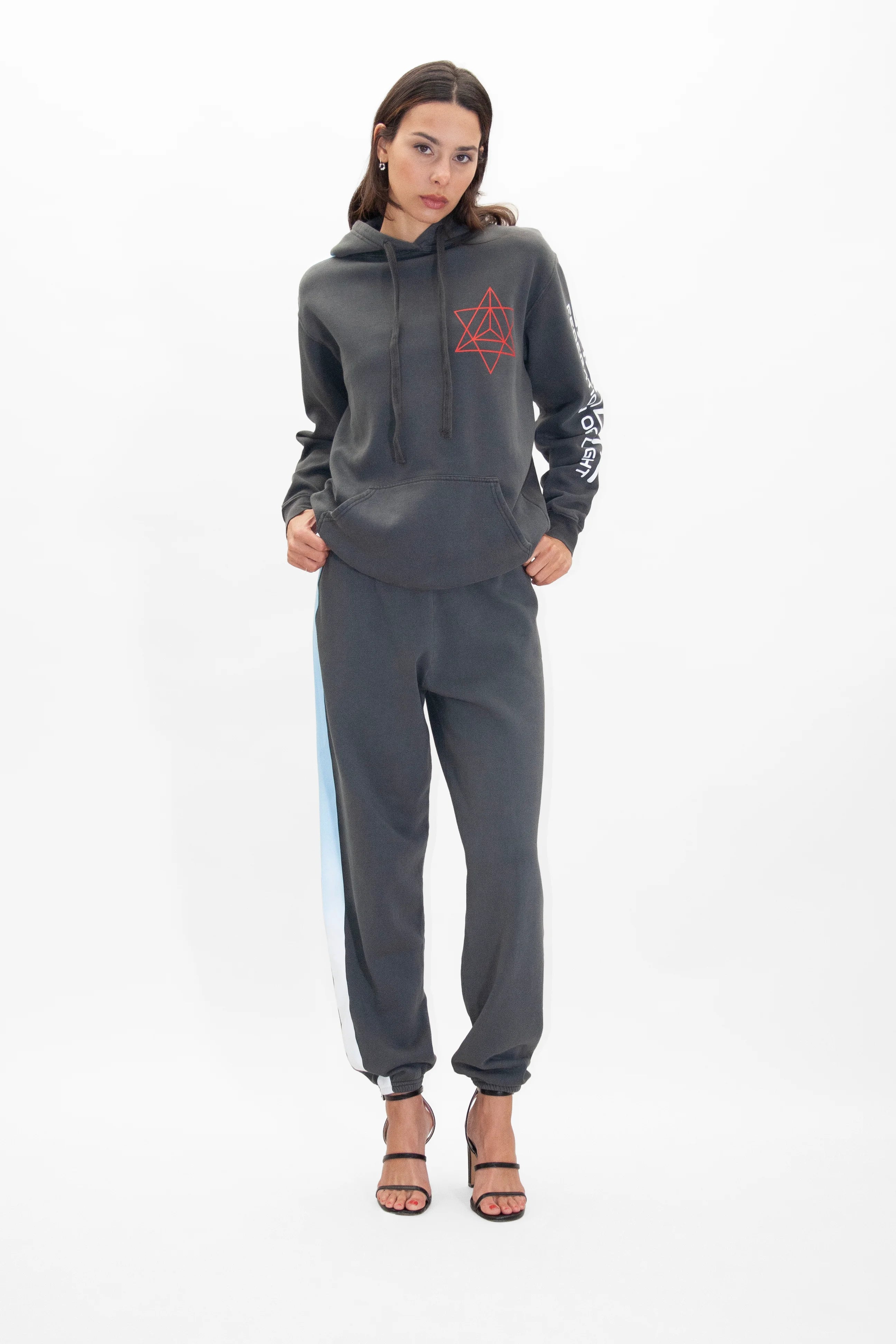 A person stands wearing the GFLApparel Merkaba Hoodie in Space Glow, matching joggers with a white-and-blue stripe on the side, and black high-heel sandals. The cotton blend outfit pops against the plain white background.