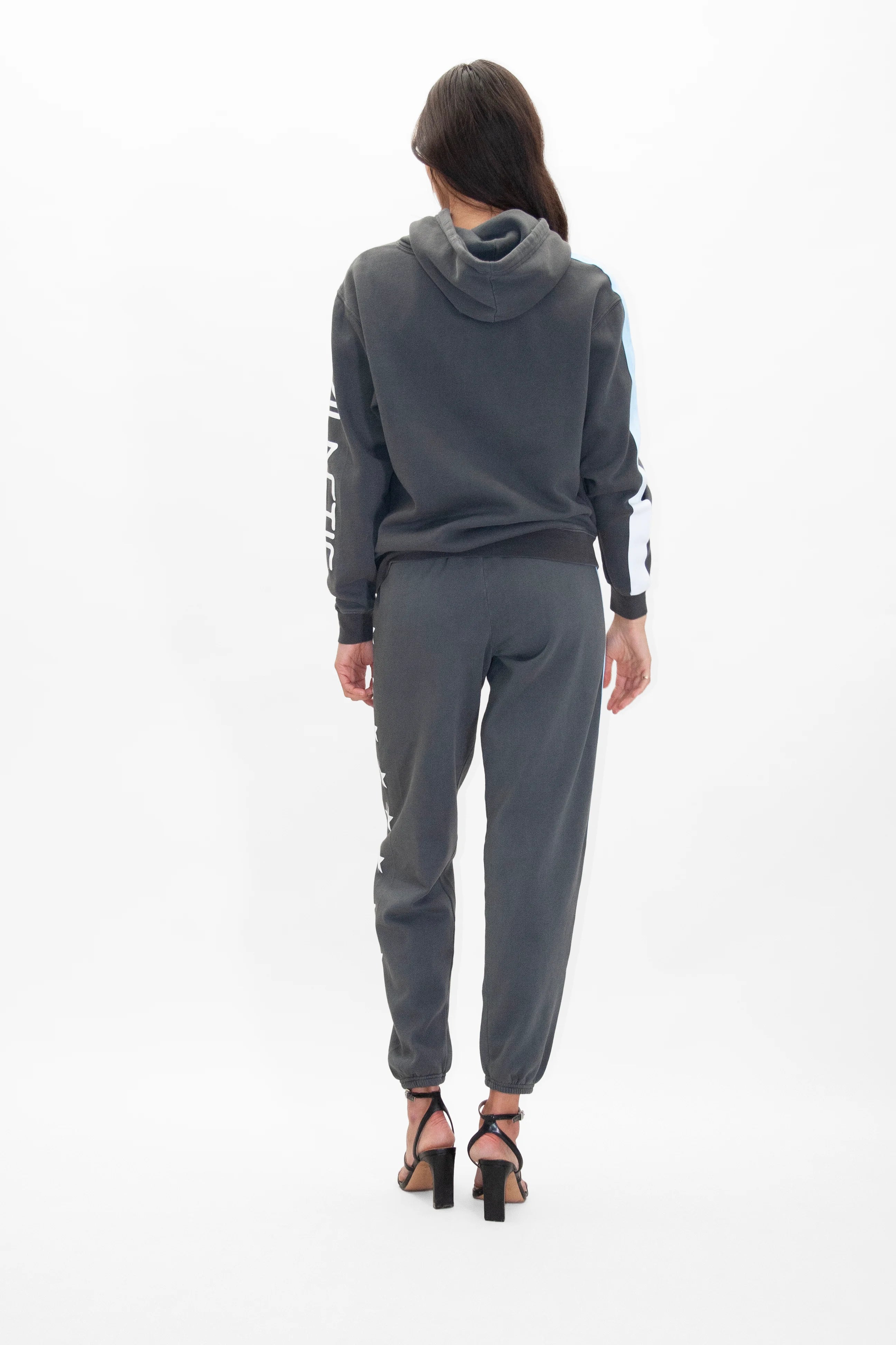 A person wearing a dark-colored GFLApparel Merkaba Hoodie in Space Glow and matching sweatpants stands facing away from the camera, adorned with black high-heeled sandals.