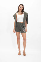 A woman stands wearing a white tank top, Merkaba Shorts in Caladan Green by GFLApparel with a white graphic on the left leg, and a matching green jacket draped over her shoulders. Her outfit, made from breathable cotton, is paired with beige open-toe sandals against a white background.
