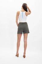 Person standing with their back to the camera, wearing a white tank top made of breathable cotton and GFLApparel Merkaba Shorts in Caladan Green, with brown heeled sandals, against a plain white background.