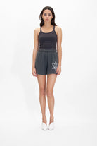 A woman with long, dark hair stands against a white background, wearing a black tank top made of breathable cotton, GFLApparel's Merkaba Shorts in Space Glow with a star design resembling sacred geometry, and white shoes.