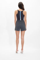 A person with dark hair is standing with their back to the camera. They are wearing a sleeveless black top, featuring a vertical blue stripe inspired by Sacred Geometry, and dark grey Merkaba Shorts in Space Glow from GFLApparel made from breathable cotton, paired with white high-heeled shoes.