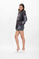 A woman is wearing a dark hoodie and matching Merkaba Shorts in Space Glow from GFLApparel, made from breathable cotton, adorned with white Sacred Geometry designs. She has long hair and stands with one leg slightly bent, wearing white high-heeled shoes.