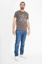 A man with a beard stands against a white background, wearing a Merkaba Tee in Earth by GFLApparel, blue jeans, and white shoes.