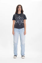 A person with long hair is standing, wearing a black GFLApparel Merkaba Tee in Space Glow featuring a sacred geometry design in breathable cotton, light blue jeans, and black sneakers. They are posing against a white background.