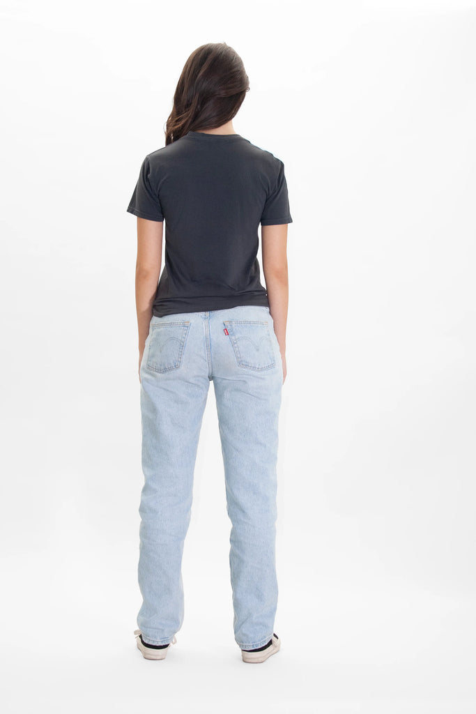 A person with long hair is standing with their back to the camera, wearing a black Merkaba Tee in Space Glow by GFLApparel, light blue jeans, and white shoes against a plain white background.