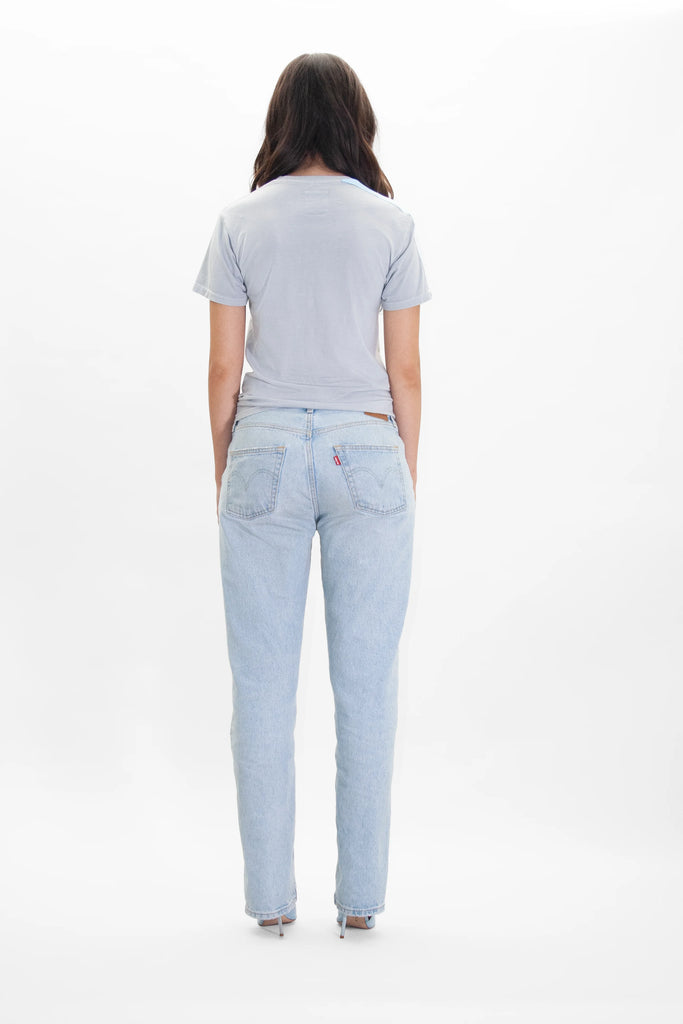 A person with long hair stands facing away, wearing a GFLApparel Moon Metatron Tee in Galactic Gray and light blue jeans against a plain white background.