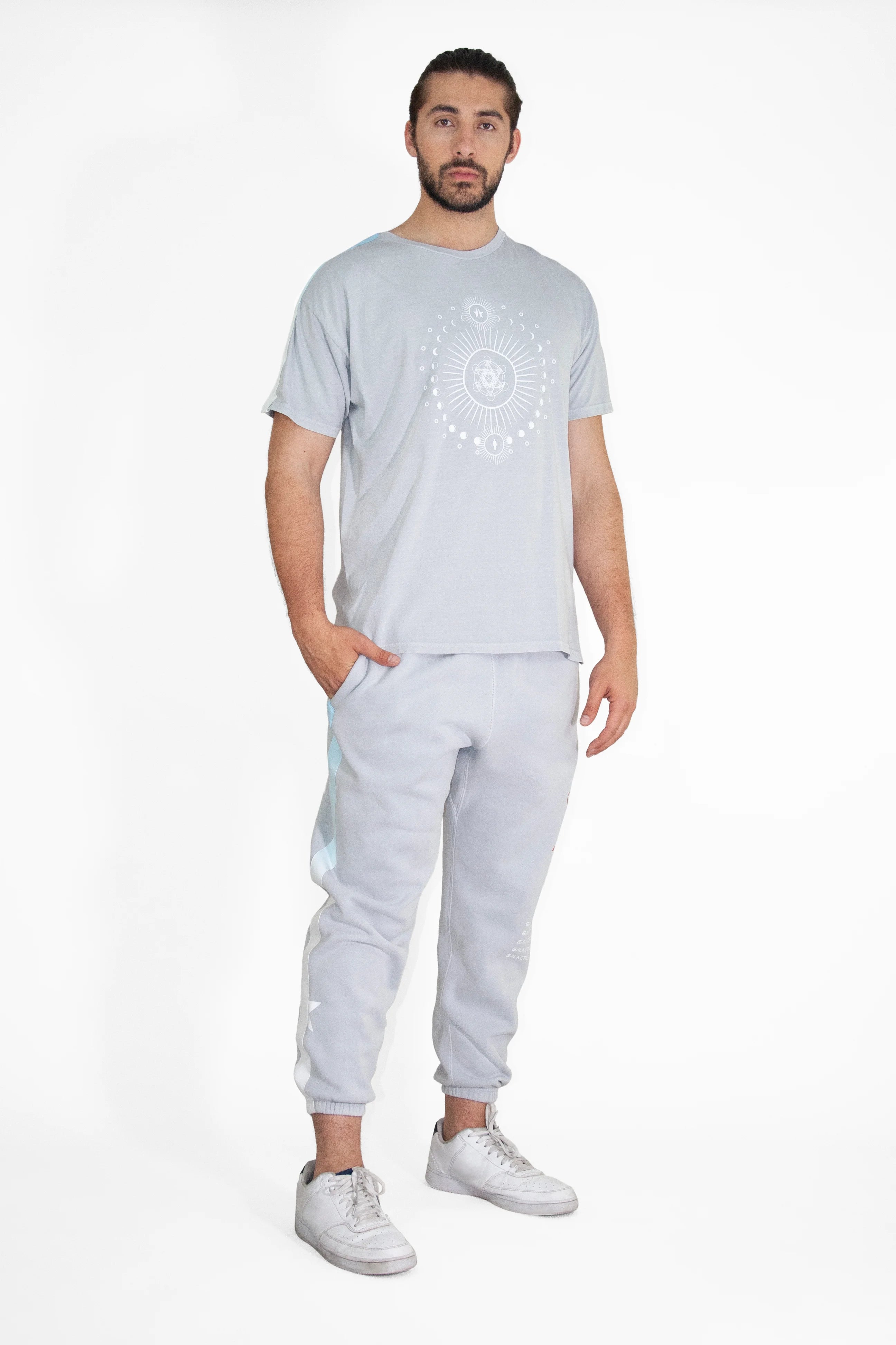 A man stands against a plain white background wearing a light grey Moon Metatron Tee in Galactic Gray from GFLApparel with a circular Sacred Geometry design, light grey sweatpants, and white sneakers. He has his right hand in his pocket.