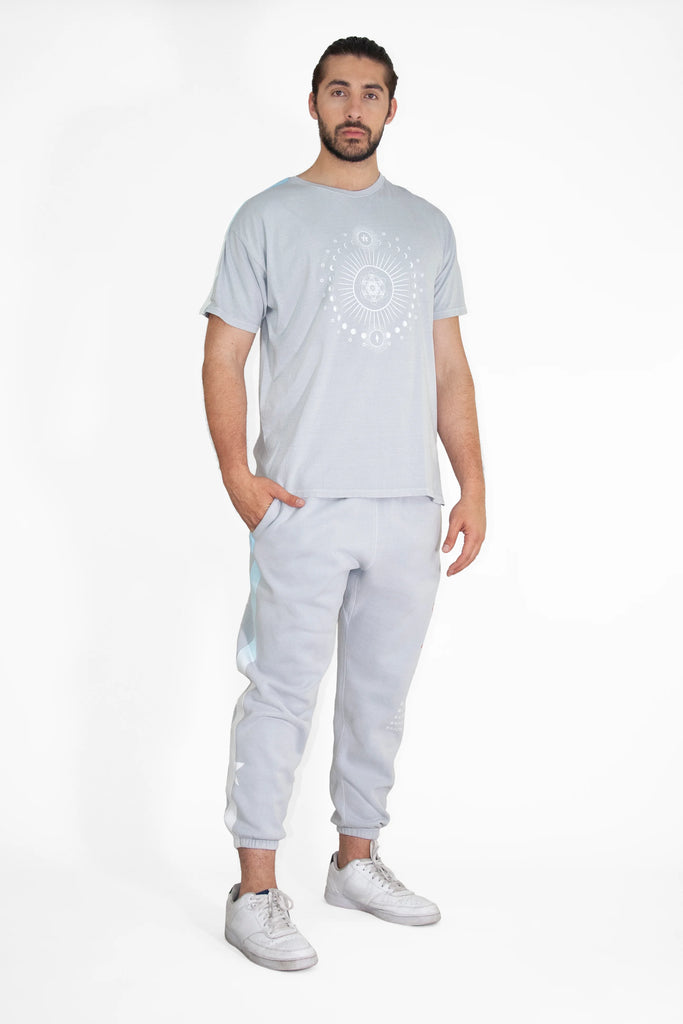 A man stands against a plain white background wearing a light grey Moon Metatron Tee in Galactic Gray from GFLApparel with a circular Sacred Geometry design, light grey sweatpants, and white sneakers. He has his right hand in his pocket.