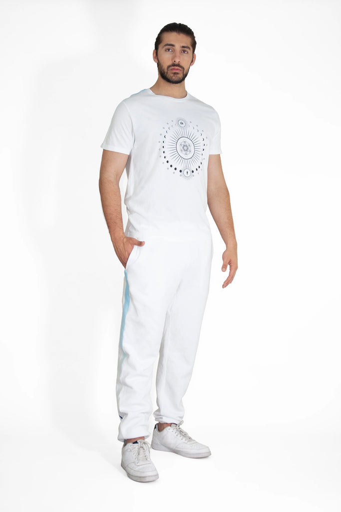 A man stands against a white background, wearing a GFLApparel Moon Metatron Tee in Lite Beam and white pants. He has his left hand in his pocket and is looking directly at the camera.
