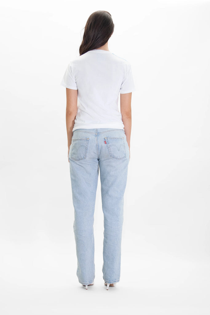 A person with long dark hair is standing with their back to the camera, wearing a GFLApparel Moon Metatron Tee in Lite Beam, light-wash jeans, and white shoes, against a plain white background.