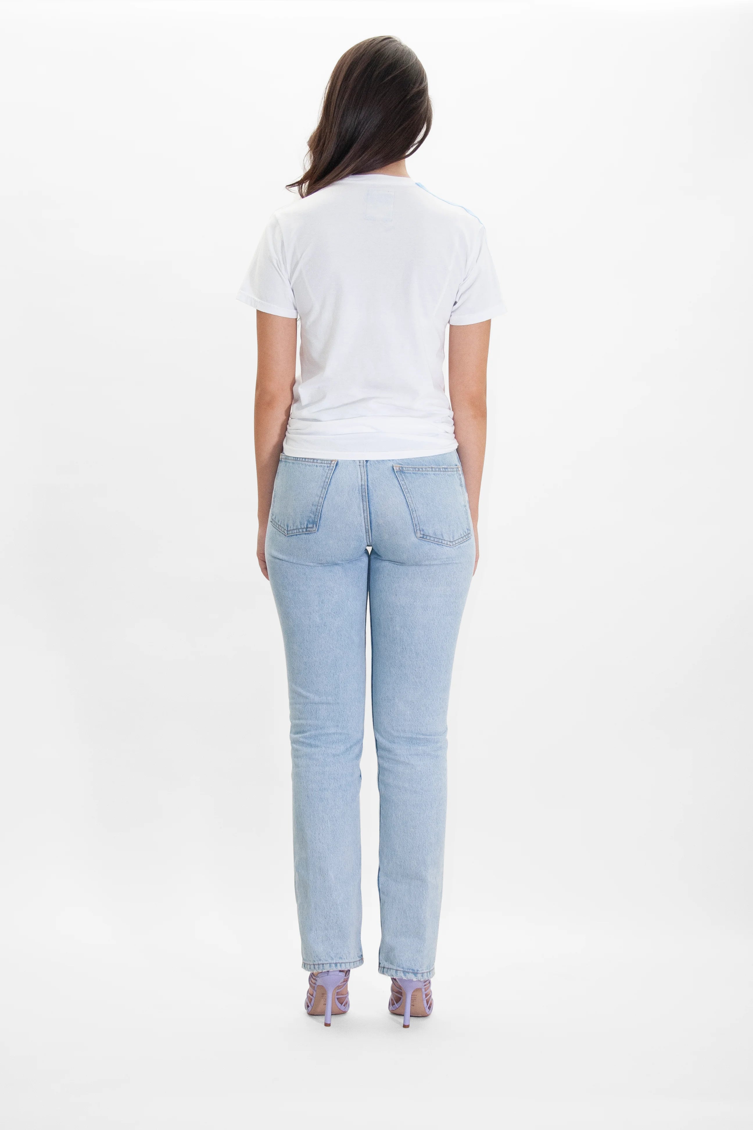 A person with long dark hair, wearing a 'More Light Tee in Lite Beam' by GFLApparel, light blue jeans, and purple heels, is standing with their back to the camera against a plain white background.