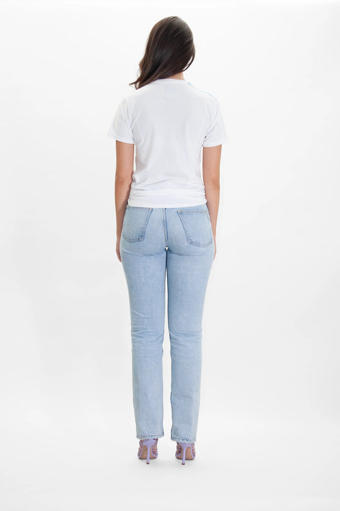 A person with long dark hair, wearing a 'More Light Tee in Lite Beam' by GFLApparel, light blue jeans, and purple heels, is standing with their back to the camera against a plain white background.