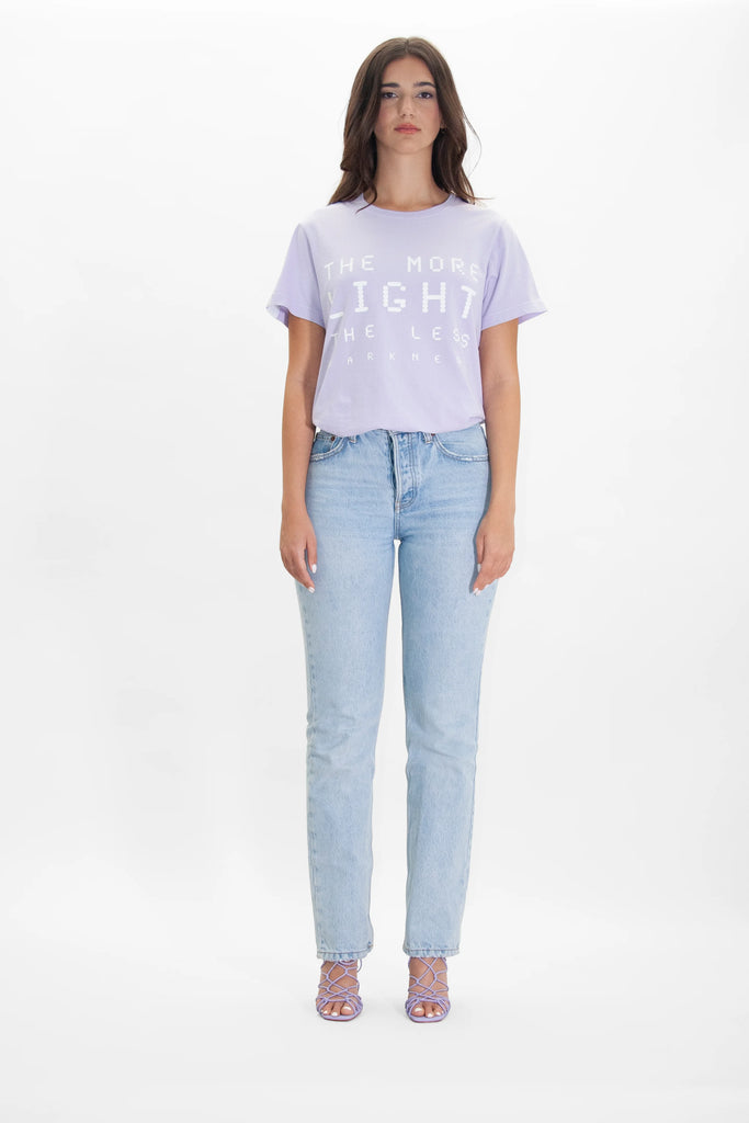 A person stands against a plain white background, wearing a light purple More Light Tee in Nebula by GFLApparel and light blue jeans, paired with comfy strappy lavender heels.