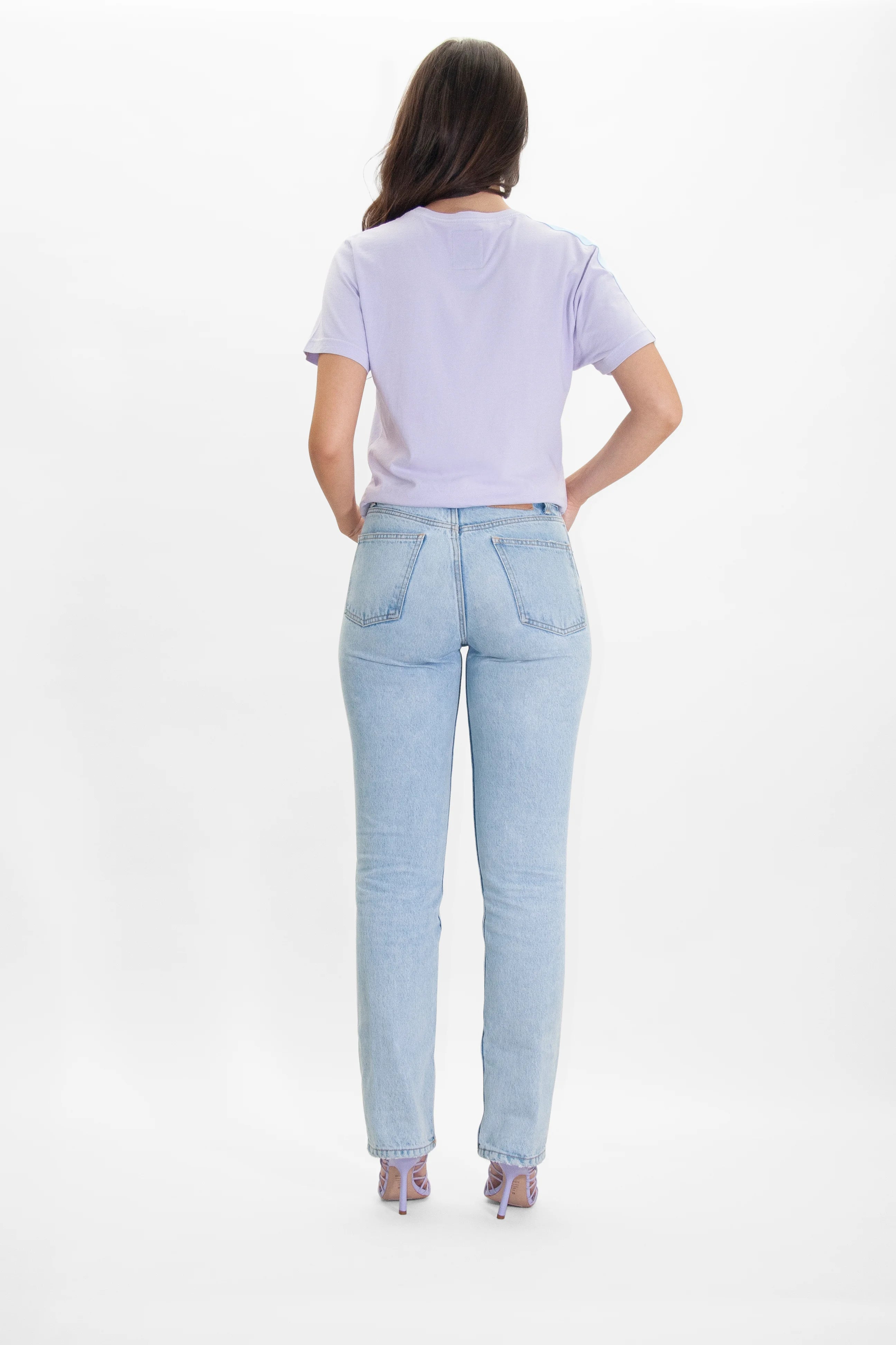 A person with long hair, wearing a comfy GFLApparel More Light Tee in Nebula, light blue jeans, and purple sandals, is standing with their back to the camera against a plain white background.