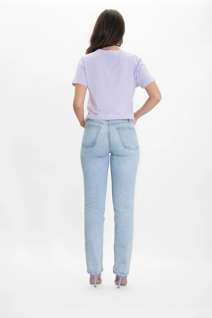 A person with long hair, wearing a comfy GFLApparel More Light Tee in Nebula, light blue jeans, and purple sandals, is standing with their back to the camera against a plain white background.