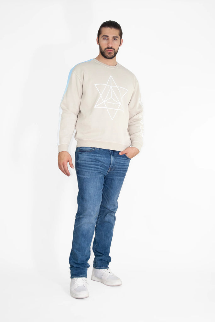 A man with short dark hair and a beard is standing against a white background. He is wearing a beige GFLApparel Merkaba Crewneck in Dune featuring a sacred geometric design, blue jeans, and white sneakers.