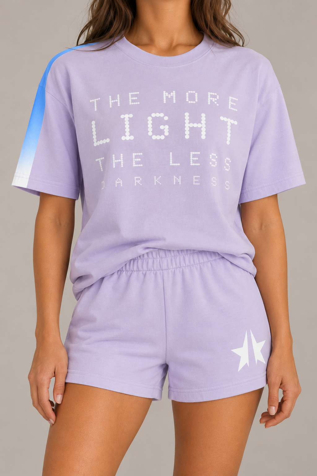 A model wearing a lavender purple matching lounge set consisting of a short-sleeve graphic t-shirt and matching shorts. The t-shirt features white text reading 'THE MORE LIGHT THE LESS DARKNESS' and a blue-to-white gradient stripe on the right sleeve.