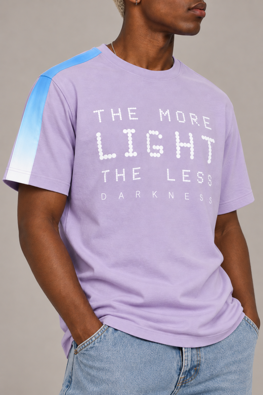 A man wearing a light purple graphic t-shirt with a blue and white gradient sleeve, featuring the text 'THE MORE LIGHT THE LESS DARKNESS' in white pixelated letters, paired with light blue jeans.