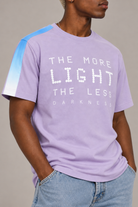 A man wearing a light purple graphic t-shirt with a blue and white gradient sleeve, featuring the text 'THE MORE LIGHT THE LESS DARKNESS' in white pixelated letters, paired with light blue jeans.