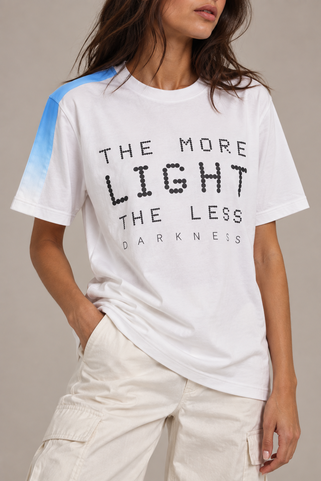 A woman wearing a white graphic T-shirt with black text reading 'THE MORE LIGHT THE LESS DARKNESS' and a blue gradient shoulder accent, paired with light cargo pants against a neutral beige background.