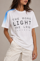 A woman wearing a white graphic T-shirt with black text reading 'THE MORE LIGHT THE LESS DARKNESS' and a blue gradient shoulder accent, paired with light cargo pants against a neutral beige background.