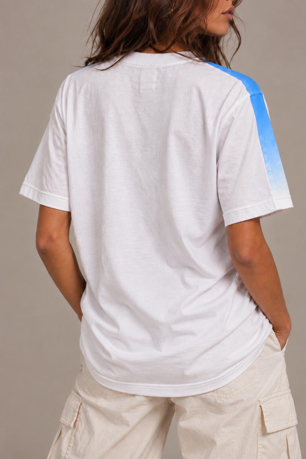 A woman wearing a white short-sleeve t-shirt with a blue gradient stripe down the right sleeve, shown from the back against a neutral studio background.