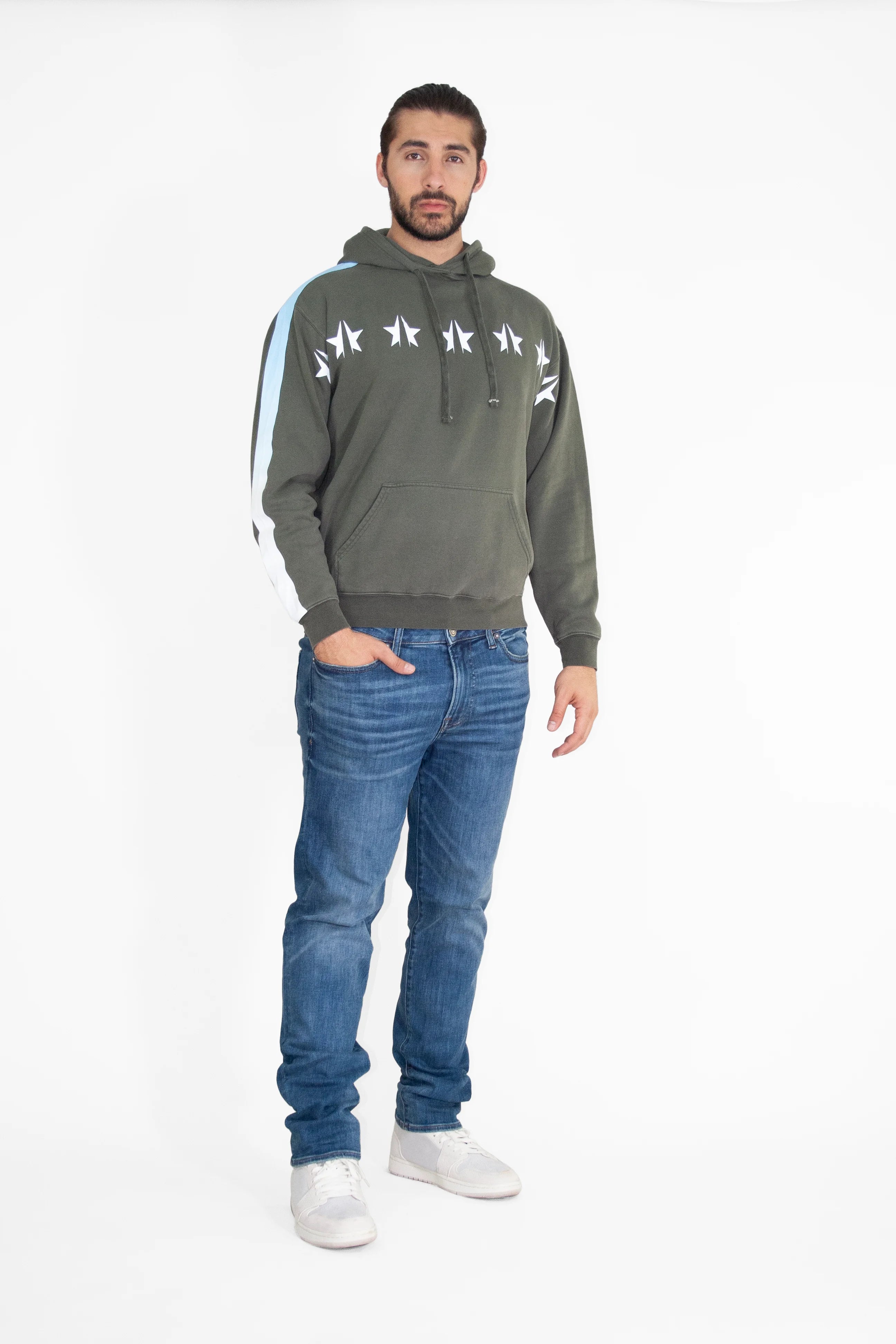 A man with dark hair and a beard stands against a white background, donning a green GFLApparel GFL Stars Hoodie in Caladan adorned with star designs, blue jeans, and white sneakers.