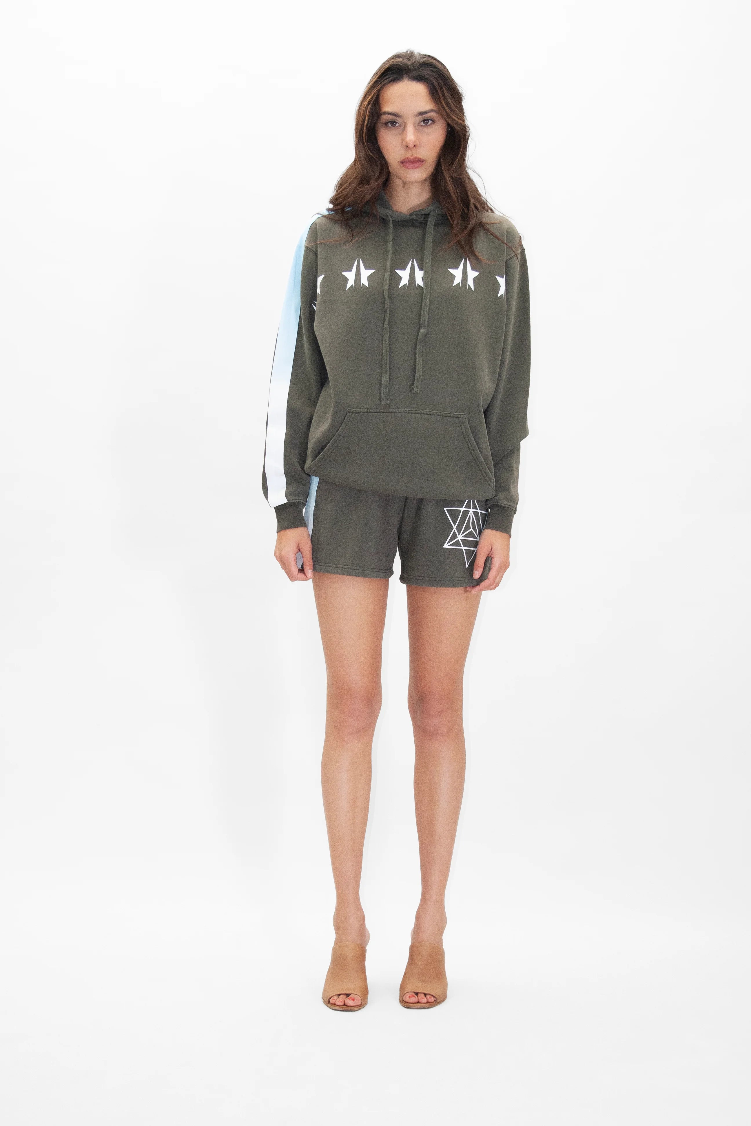 A person standing and wearing a green GFLApparel GFL Stars Hoodie in Caladan with star designs and matching green shorts. The outfit includes a white and blue stripe on one sleeve and geometric patterns on the shorts.