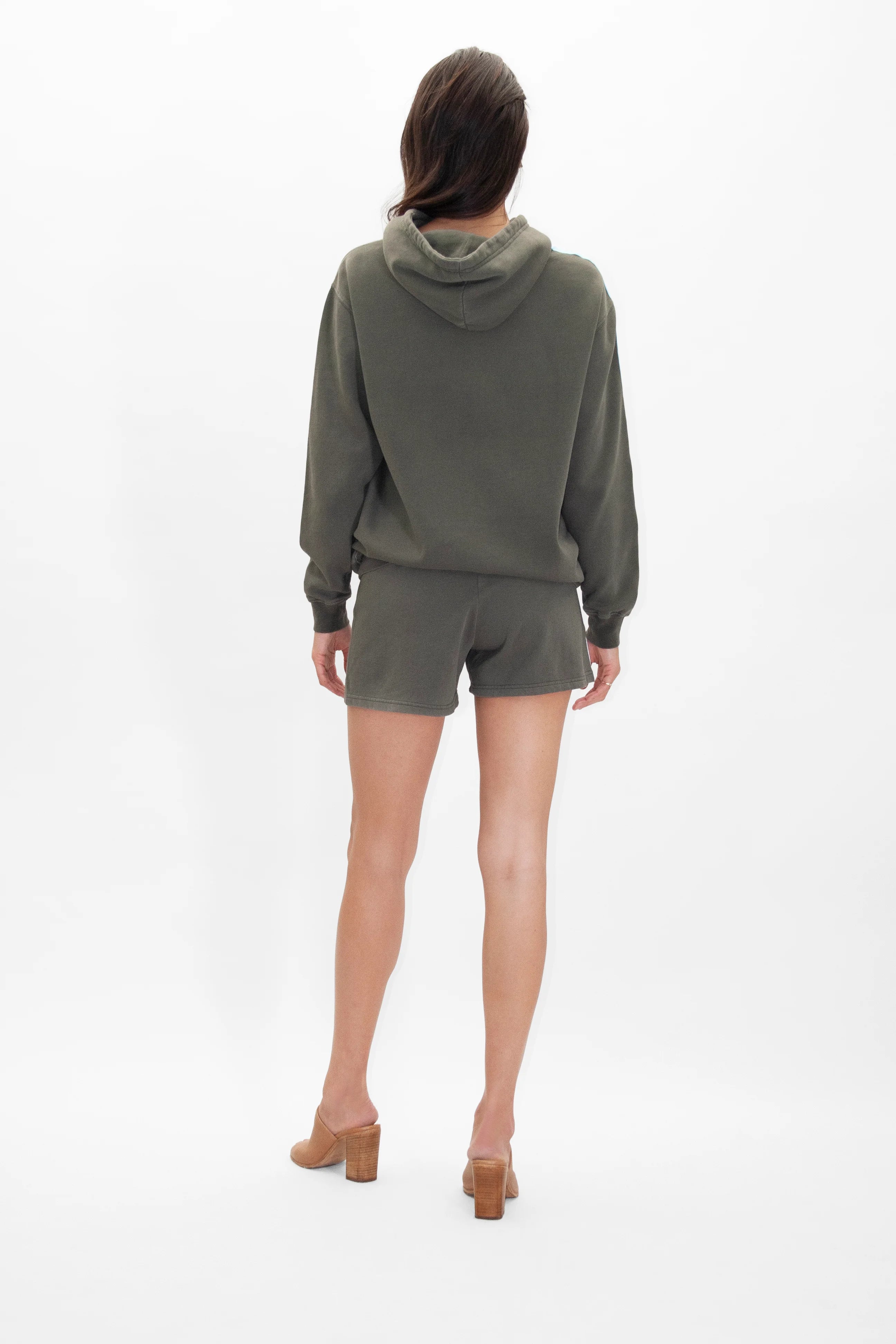 A person is standing with their back to the camera, donning a GFLApparel GFL Stars Hoodie in Caladan and matching green shorts, along with brown high-heeled shoes, against a plain white background.