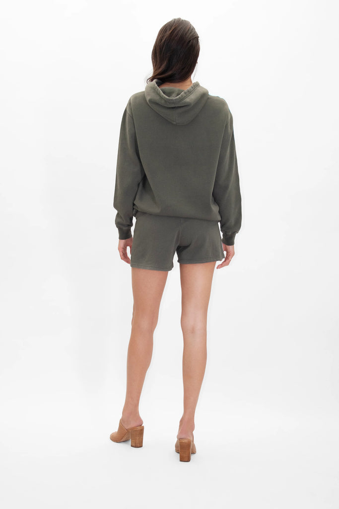 A person is standing with their back to the camera, donning a GFLApparel GFL Stars Hoodie in Caladan and matching green shorts, along with brown high-heeled shoes, against a plain white background.