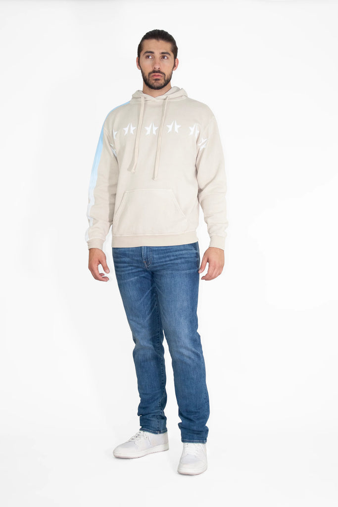 A man wearing a beige GFLApparel GFL Stars Hoodie in Dune, blue jeans, and white sneakers stands against a plain white background.