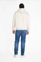 A person with tied-back hair is standing with their back to the camera, wearing a GFLApparel GFL Stars Hoodie in Dune, blue jeans, and white shoes. The background is plain white.