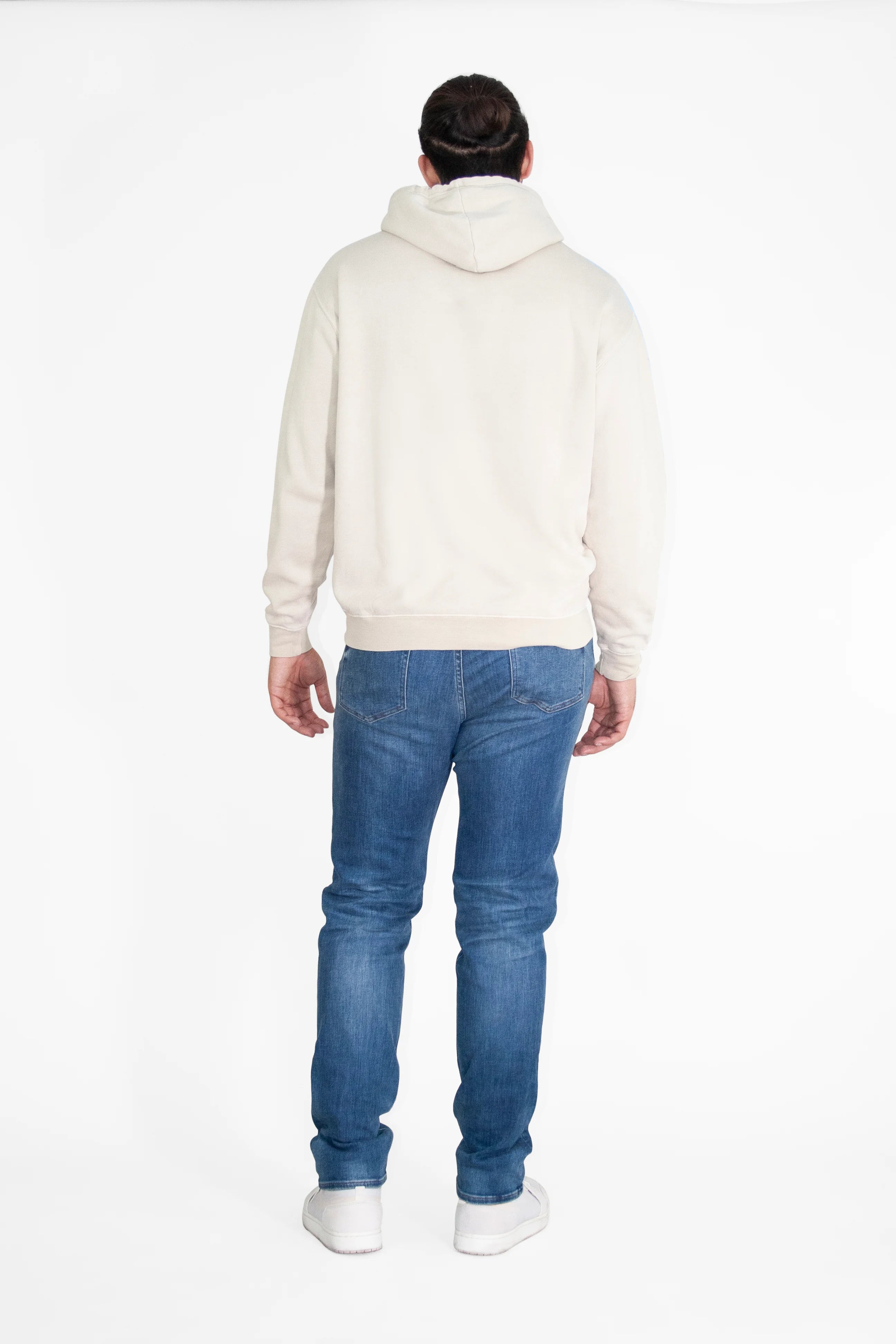 A person with tied-back hair is standing with their back to the camera, wearing a GFLApparel GFL Stars Hoodie in Dune, blue jeans, and white shoes. The background is plain white.