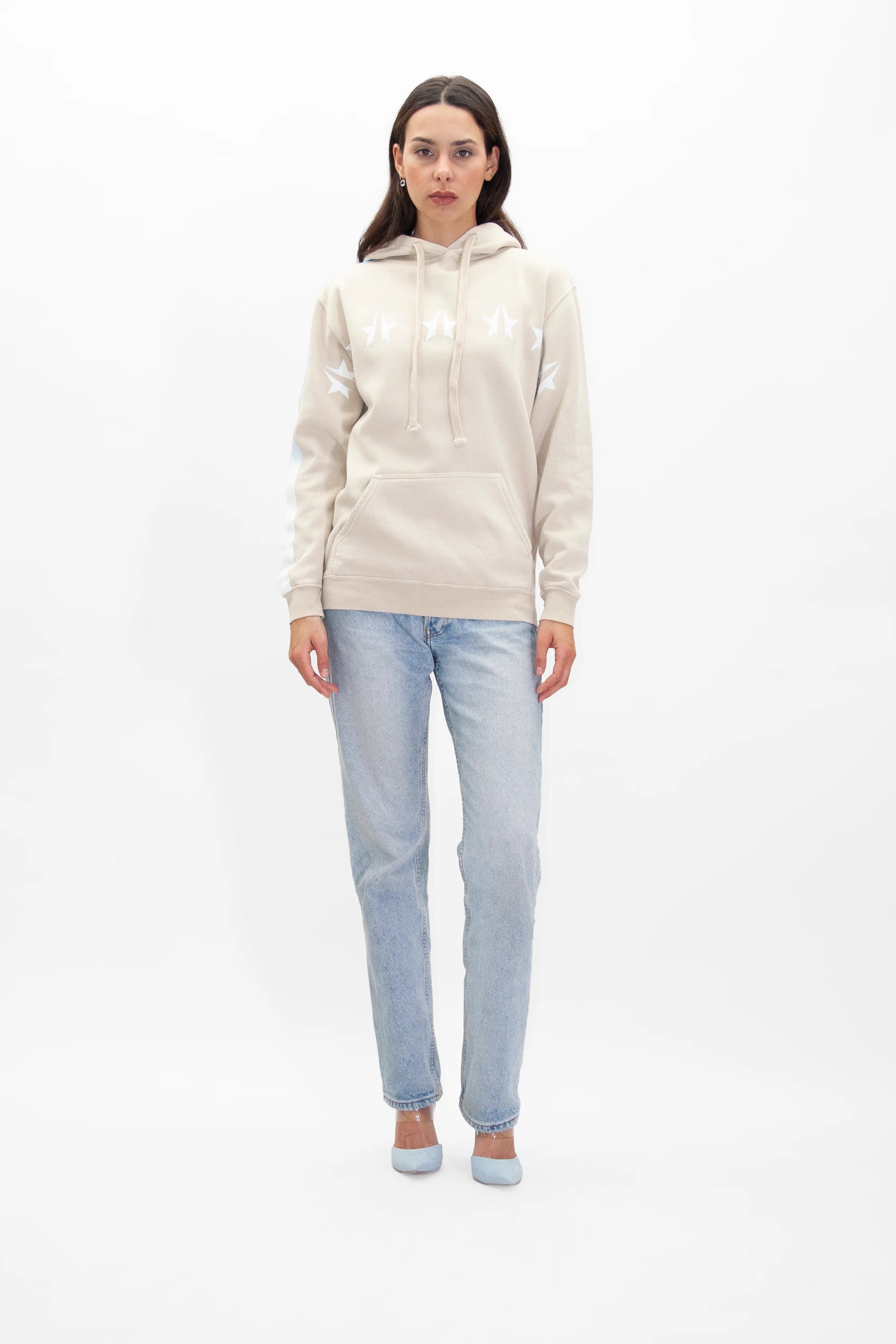 A person stands against a white background wearing a light beige GFLApparel GFL Stars Hoodie in Dune with white star designs, light blue jeans, and matching light blue shoes.