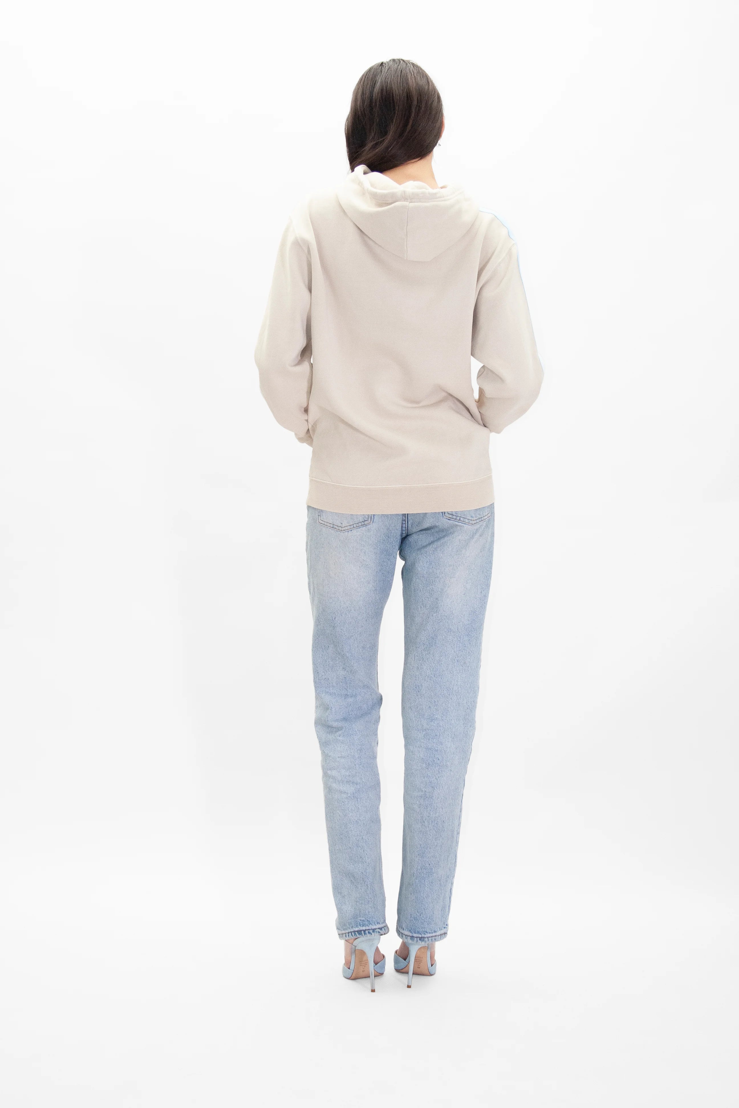 A person stands facing away, wearing a GFLApparel GFL Stars Hoodie in Dune, light blue jeans, and light-colored high-heeled sandals against a plain white background.