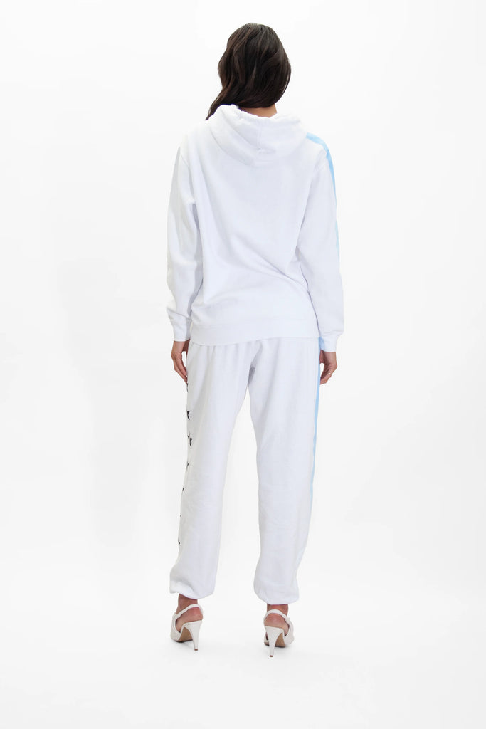 A person with long hair stands facing away, wearing a GFLApparel GFL Stars Hoodie in Lite Beam and matching sweatpants made of soft, breathable cotton with small star details on the side, and white high-heeled shoes. The background is plain white.