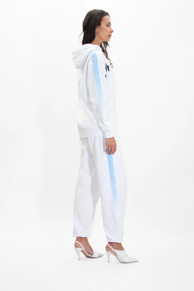 A woman stands in profile wearing a white GFLApparel GFL Stars Hoodie in Lite Beam and matching sweatpants with light blue stripes, both made from soft breathable cotton. She is also wearing white high-heeled shoes. The background is plain white.