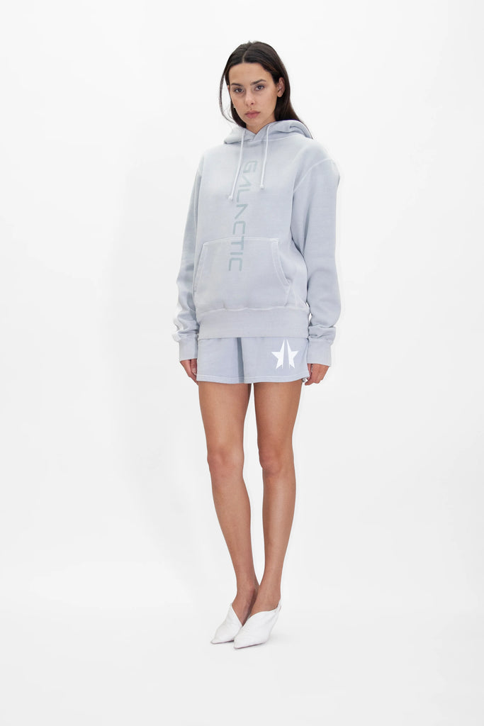 A person is standing and wearing a light gray hoodie with the word "GALACTIC" in bold print and matching GFLApparel Star Shorts in Galactic Gray. The hoodie has a hood with drawstrings, and the person is wearing white shoes.